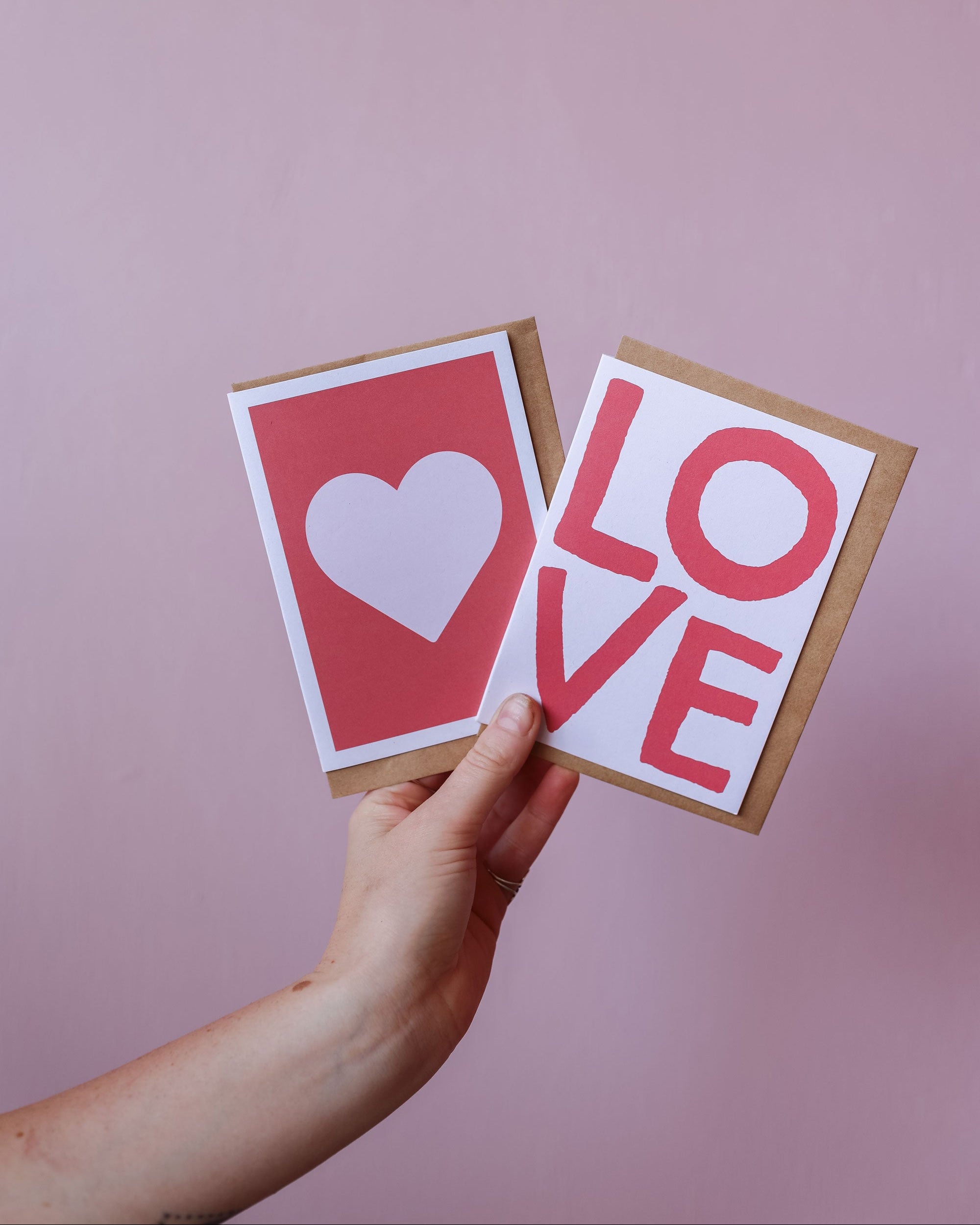 Hand holding two greeting cards with heart and 'LOVE' text against a pink background