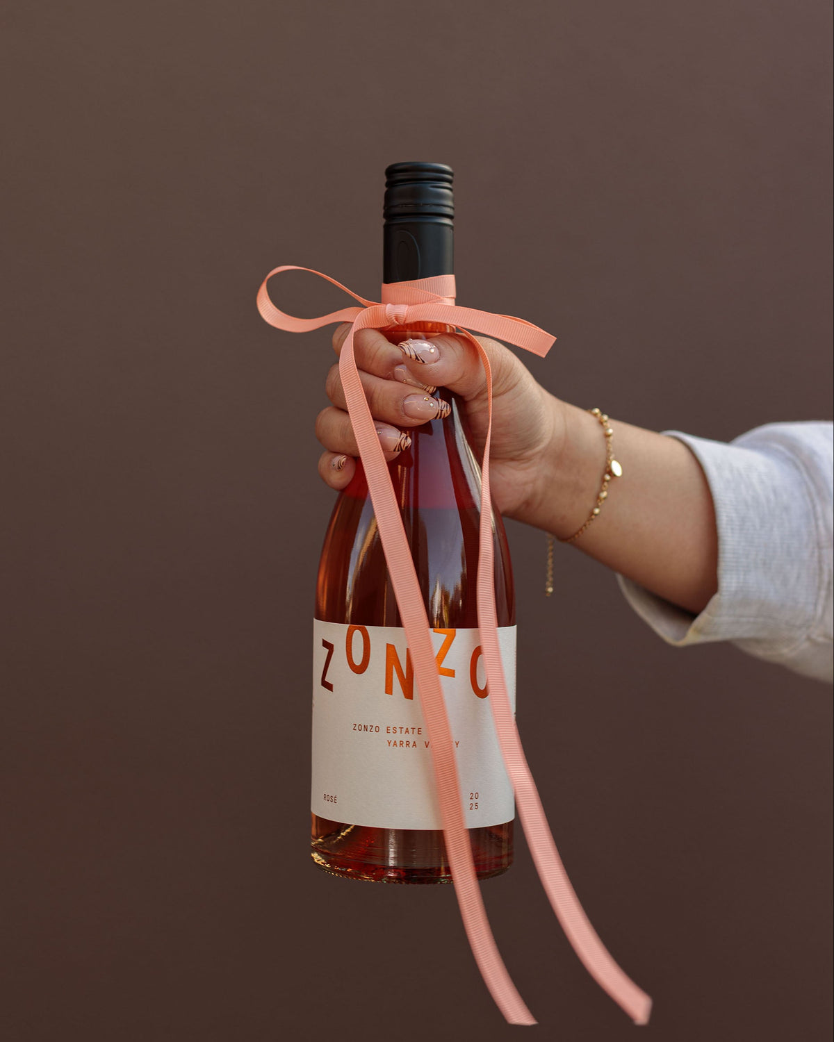 A hand holding a bottle of Zonzo rosé wine with a peach ribbon tied around the neck, set against a solid dark brown background.